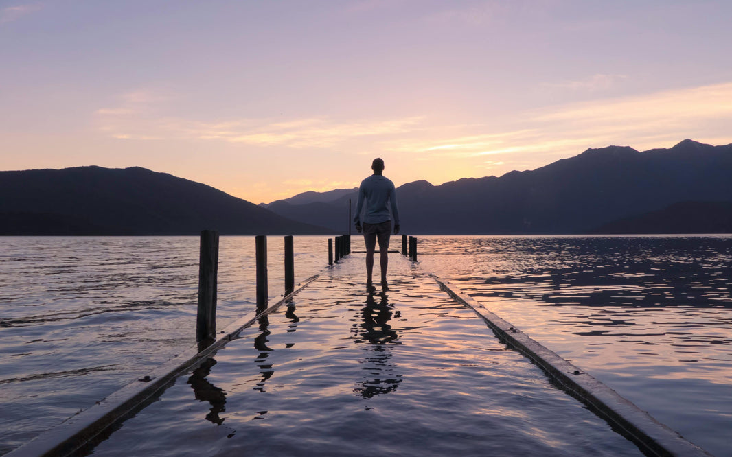 Person standing alone on a dock at sunset overlooking calm mountain waters, evoking solitude, freedom, reflection, and the spirit of exploration.