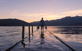Person standing alone on a dock at sunset overlooking calm mountain waters, evoking solitude, freedom, reflection, and the spirit of exploration.